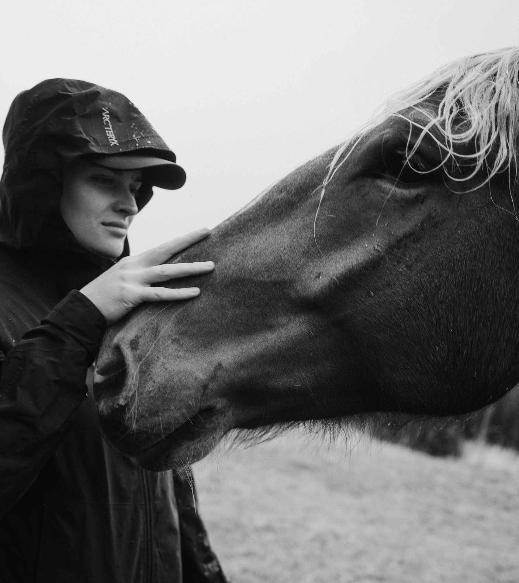 Black and white photograph of Vali Höll stroking a horse while wearing a hooded raincoat.